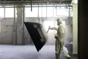 view of a worker wearing a full white protective suit and breathing mask, sand blasting a metal crate hung from a metal beam in the ceiling of an industrial hall